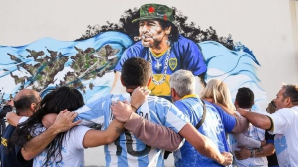 Maradona con la camiseta de Boca, fumando un habano y su gorra revolucionaria, junto a una bandera de Argentina y las Islas Malvinas.